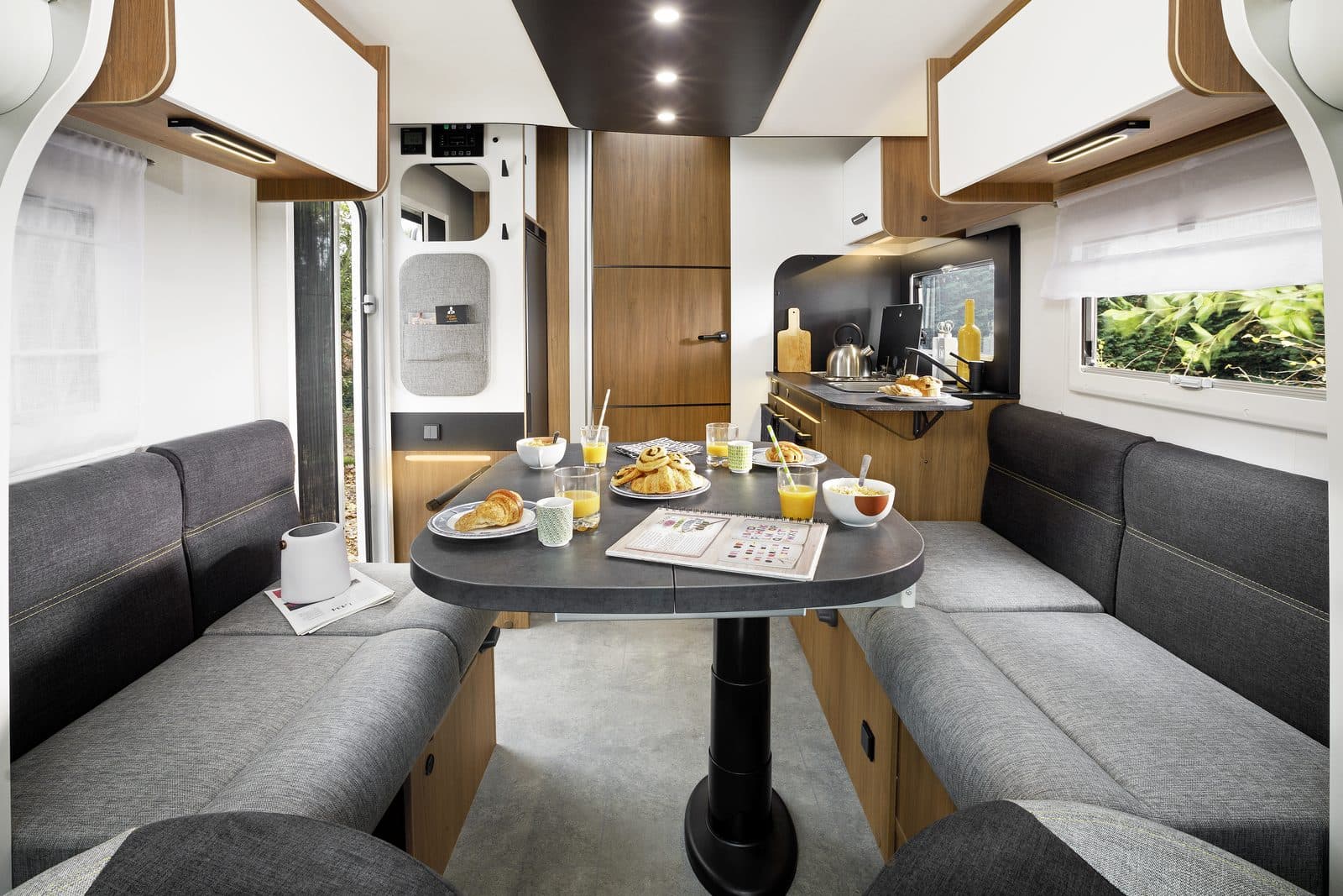 Salon d’un profilé Challenger 317 : dînette face-à-face avec banquettes grises, table centrale dressée, cuisine et grandes baies vitrées, ambiance lumineuse bois clair