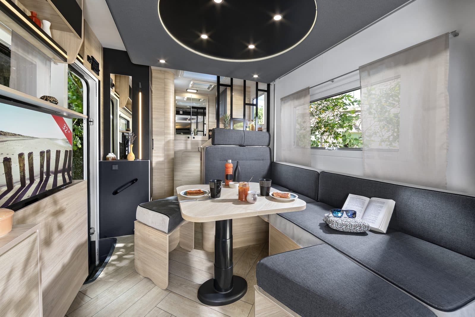Salon d’un profilé Chausson 660 : dînette face-à-face avec banquettes grises, table centrale, cuisine en fond et grandes baies vitrées, ambiance lumineuse bois clair