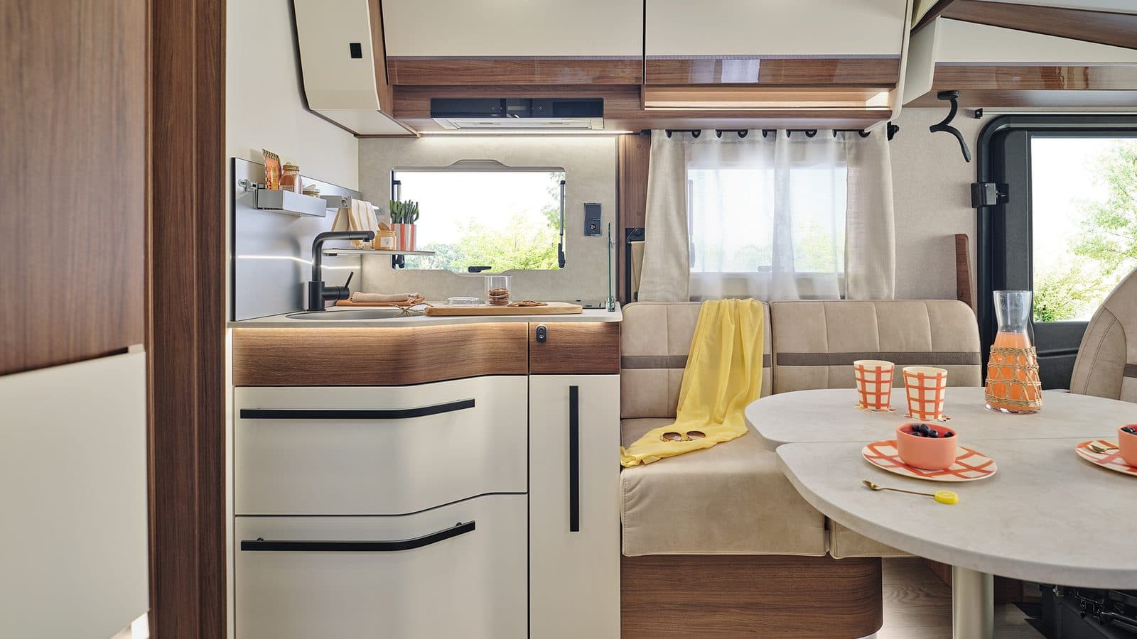 Intérieur d’un Fleurette Discover 74 LJG : cuisine aux tiroirs blancs et bois noyer, banquette beige avec plaid jaune et table de salon dressée, vers la cabine