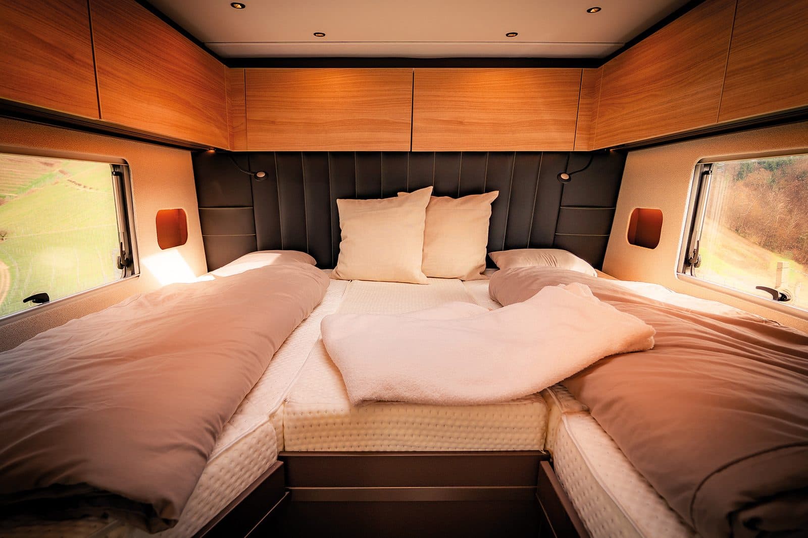 Chambre arrière d’un Bürstner Signature en configuration lits jumeaux réunis par un matelas d’appoint, têtes de lit capitonnées sombres, rangements en bois clair et grandes baies latérales donnant sur le vignoble