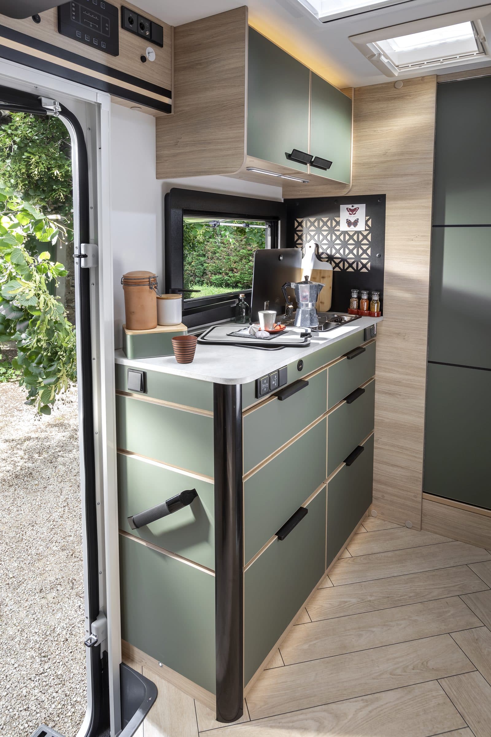 Cuisine du Chausson X650 Exclusive Line : meubles à façades vert amande et liserés bois, plan de travail blanc compact avec cafetière italienne, fenêtre et crédence noire décorée