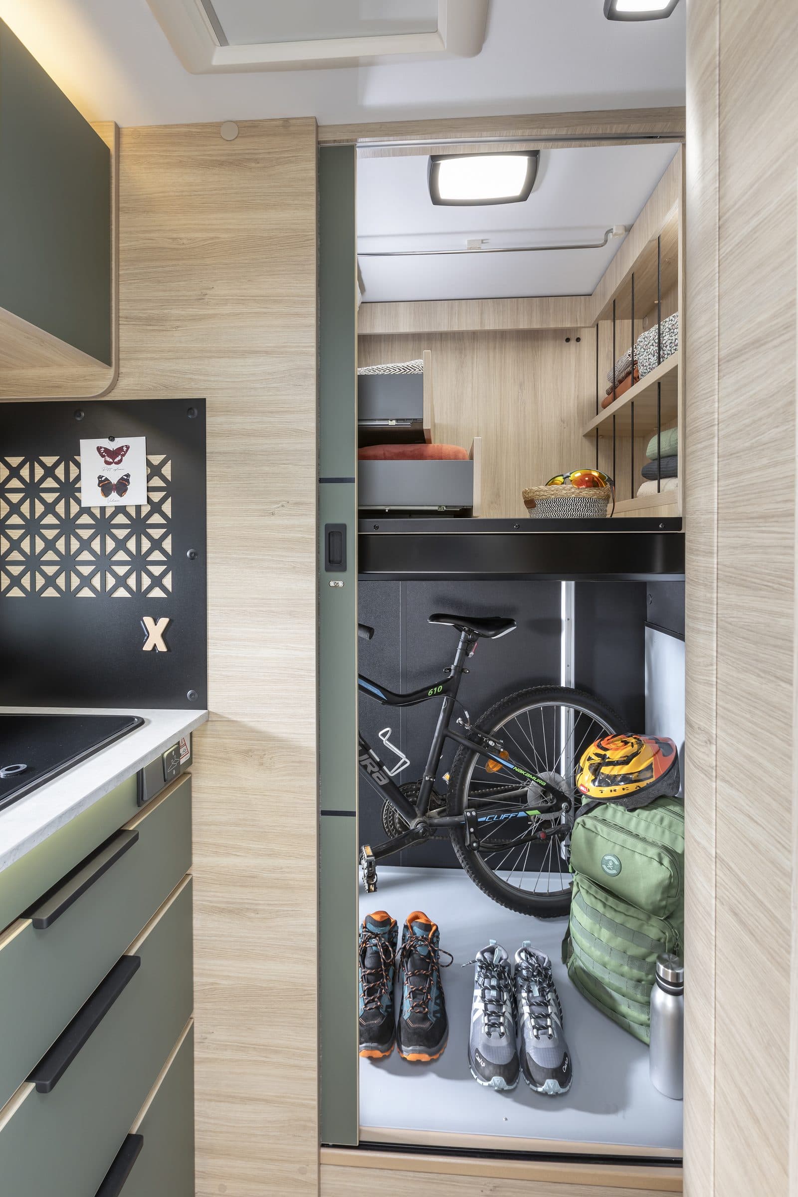 Smart Dressing Room du Chausson X650 vu depuis la cuisine : espace arrière avec un VTT chargé au sol, sac à dos et chaussures de randonnée, lit escamoté au-dessus