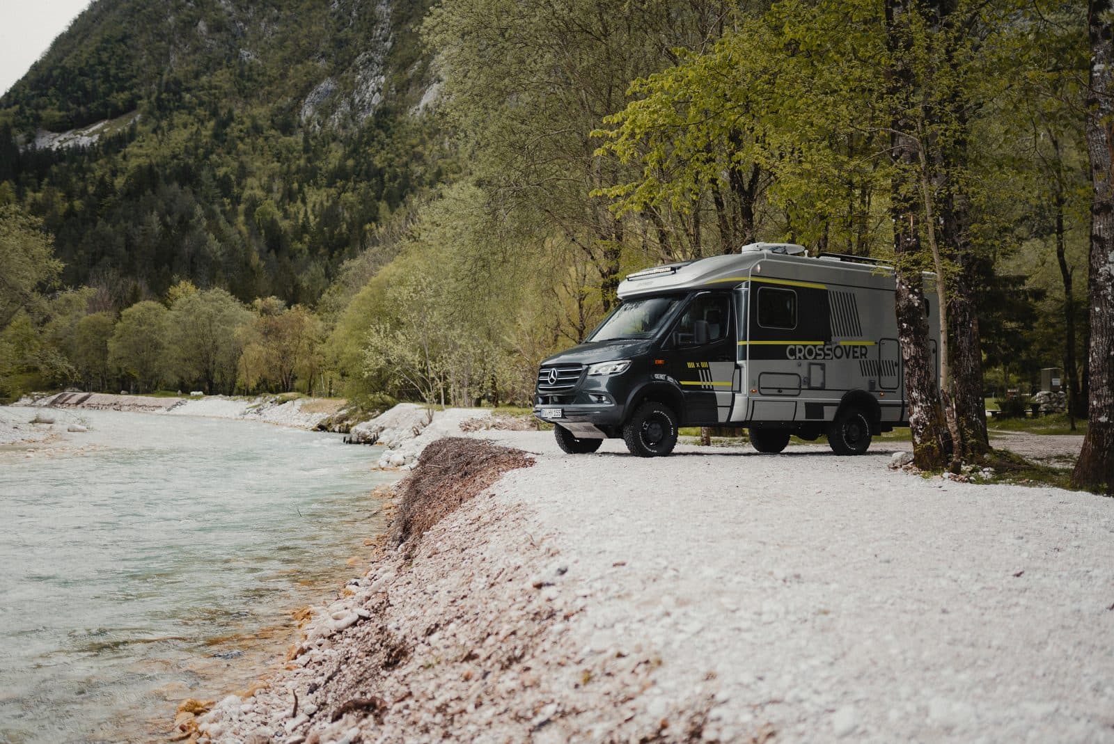 Hymer ML-T 570 CrossOver sur base Mercedes Sprinter 4x4, cabine grise et pneus tout-terrain, stationné au bord d’une rivière turquoise de montagne dans une vallée boisée