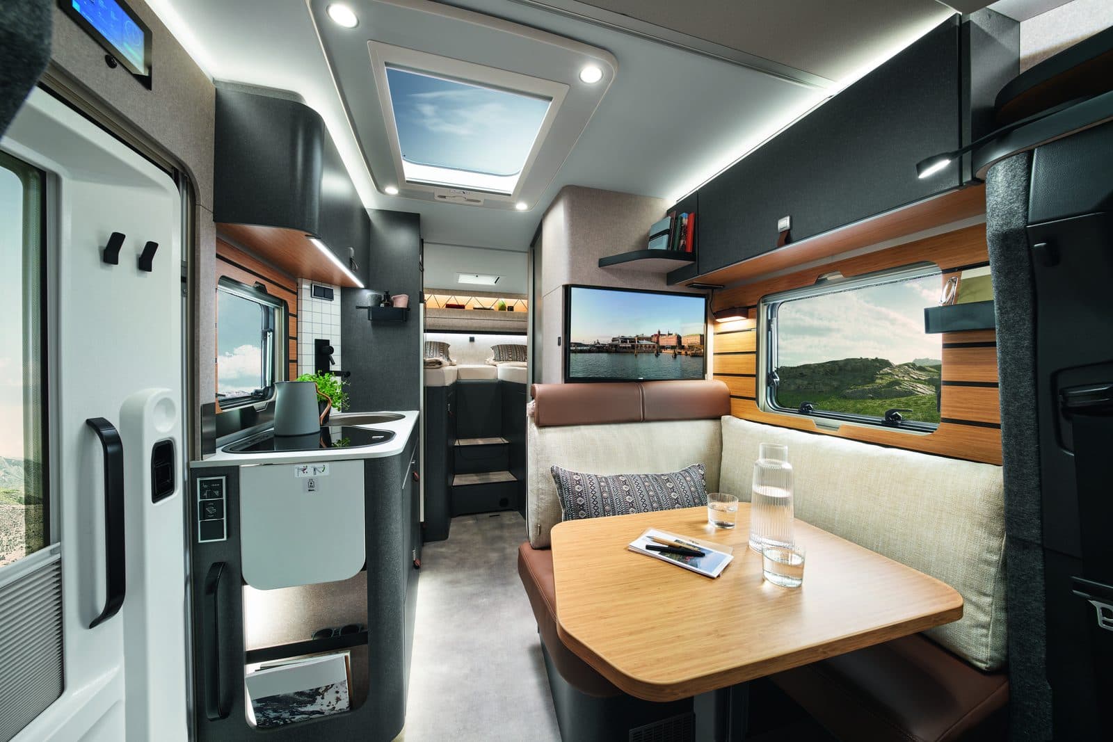 Intérieur lumineux du Hymer ML-T 570 : dînette avec sièges pivotants et coussins clairs, table en bois, grande baie latérale et lanterneau panoramique, cuisine compacte et lits jumeaux visibles à l’arrière